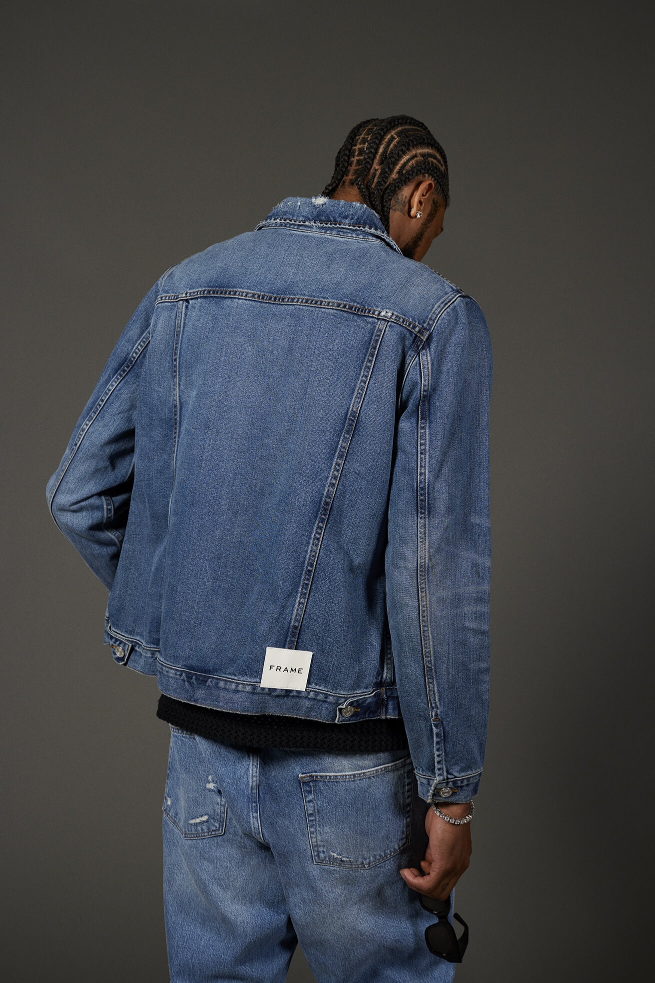 A person with braided hair poses facing away wearing a denim jacket and matching jeans against a plain gray backdrop in a studio setting.