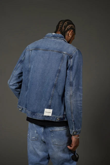 A person with braided hair poses facing away wearing a denim jacket and matching jeans against a plain gray backdrop in a studio setting.