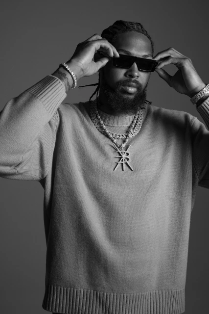 Black and white portrait of an adult man with a beard wearing sunglasses and jewelry, posing in front of a neutral background.
