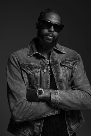 A portrait in black and white of a stylish adult male with a beard, sunglasses, and a denim jacket posing against a dark background.