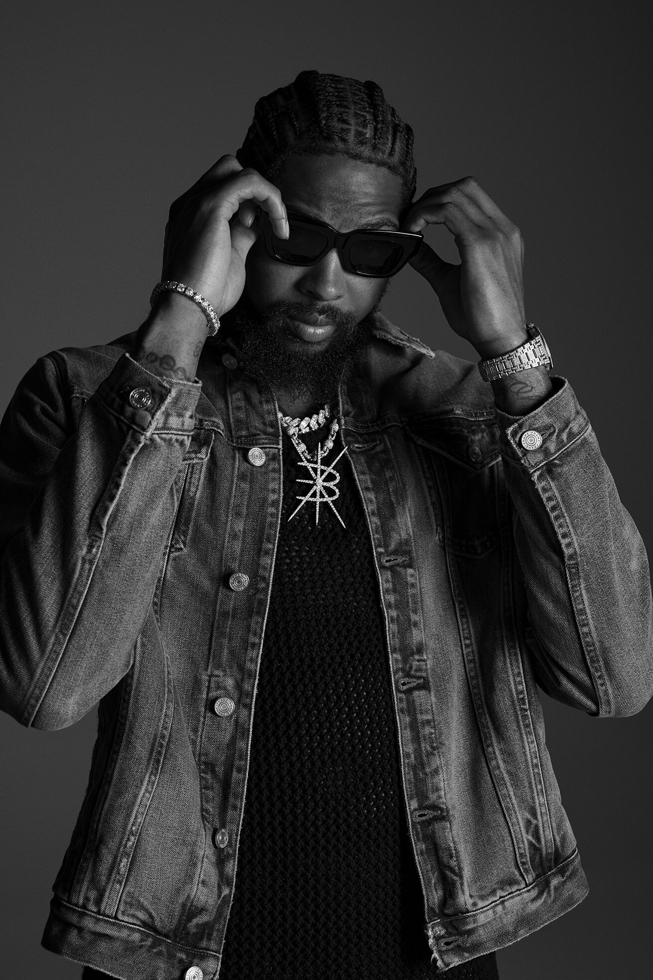 Monochrome portrait of an adult male with a beard wearing sunglasses, a denim jacket, and jewelry against a grey background.