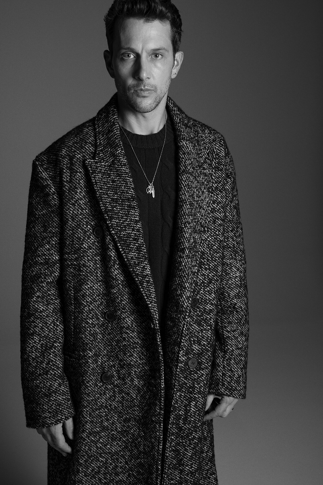 Black and white portrait of a man in a patterned coat, wearing a pendant, against a gray background in a studio setting.