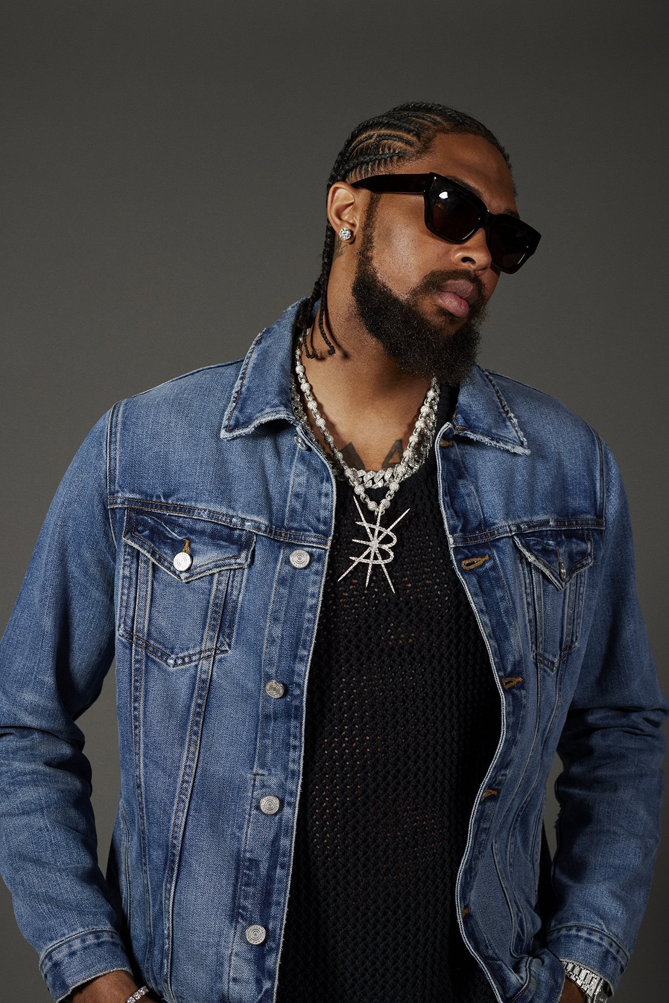 Portrait of a bearded adult male in a denim jacket and sunglasses, wearing jewelry, against a neutral grey studio backdrop.