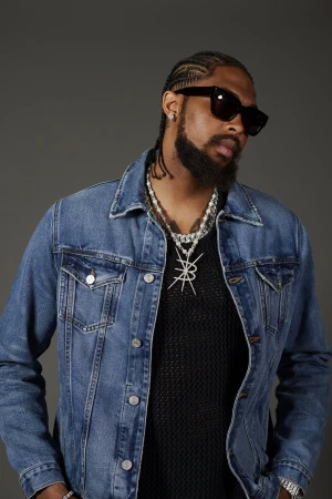Portrait of a bearded adult male in a denim jacket and sunglasses, wearing jewelry, against a neutral grey studio backdrop.