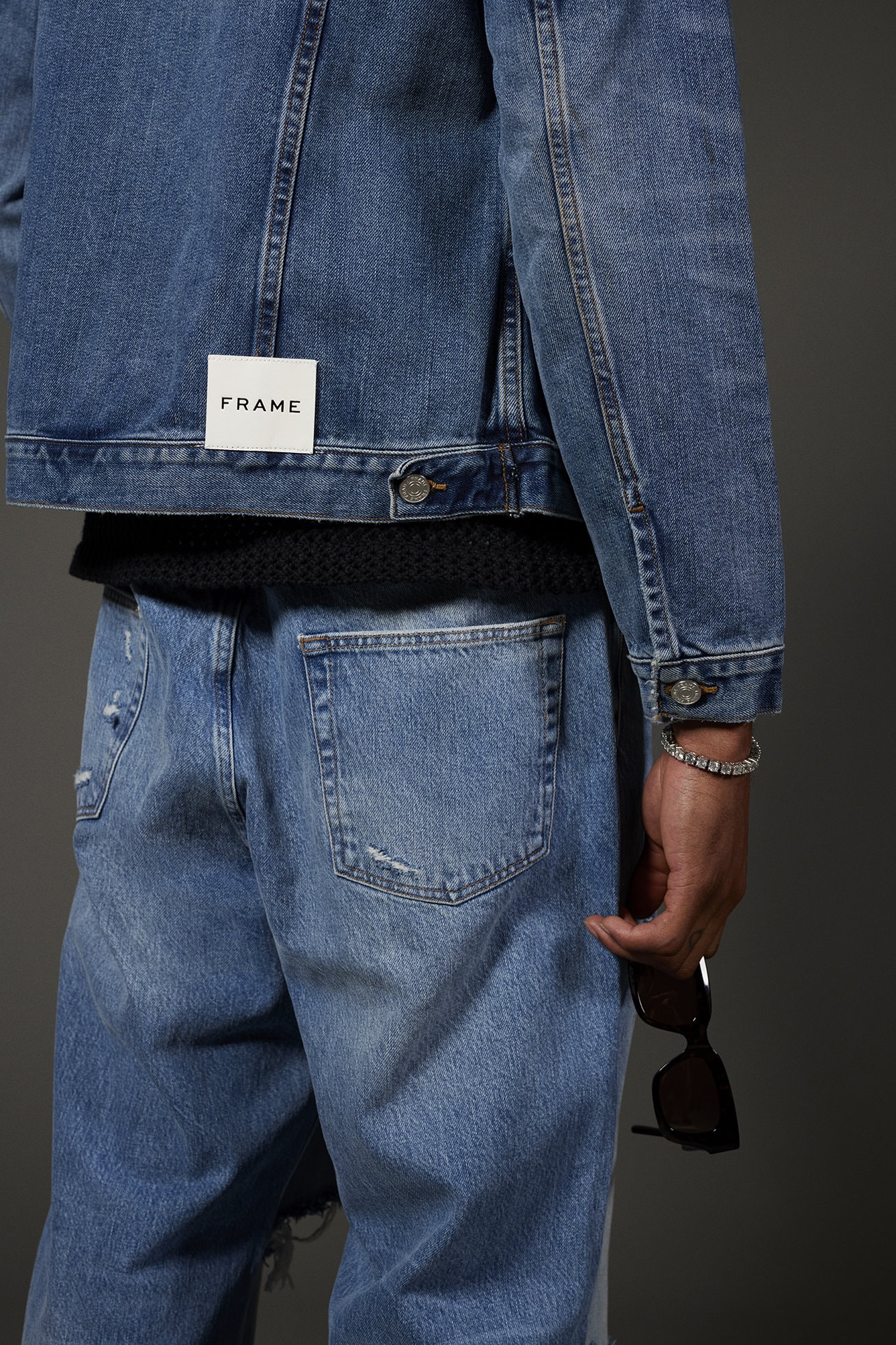 Back view of a person wearing a FRAME denim jacket and light blue jeans, holding sunglasses in their hand, studio shot on gray.