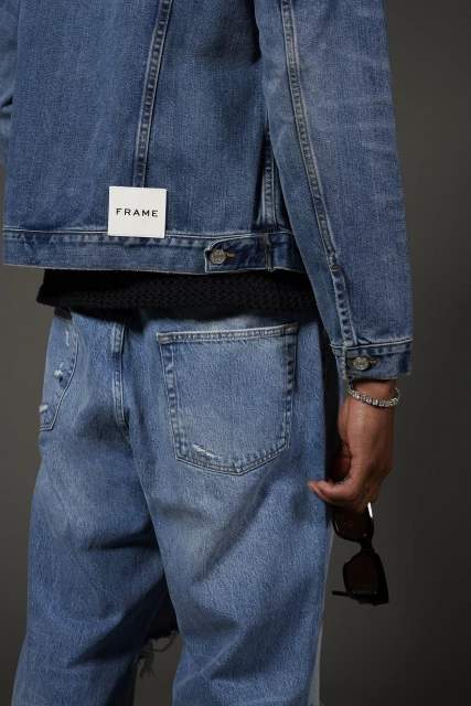 Back view of a person wearing a FRAME denim jacket and light blue jeans, holding sunglasses in their hand, studio shot on gray.