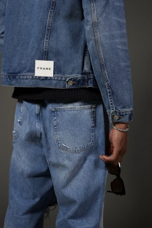 Back view of a person wearing a FRAME denim jacket and light blue jeans, holding sunglasses in their hand, studio shot on gray.