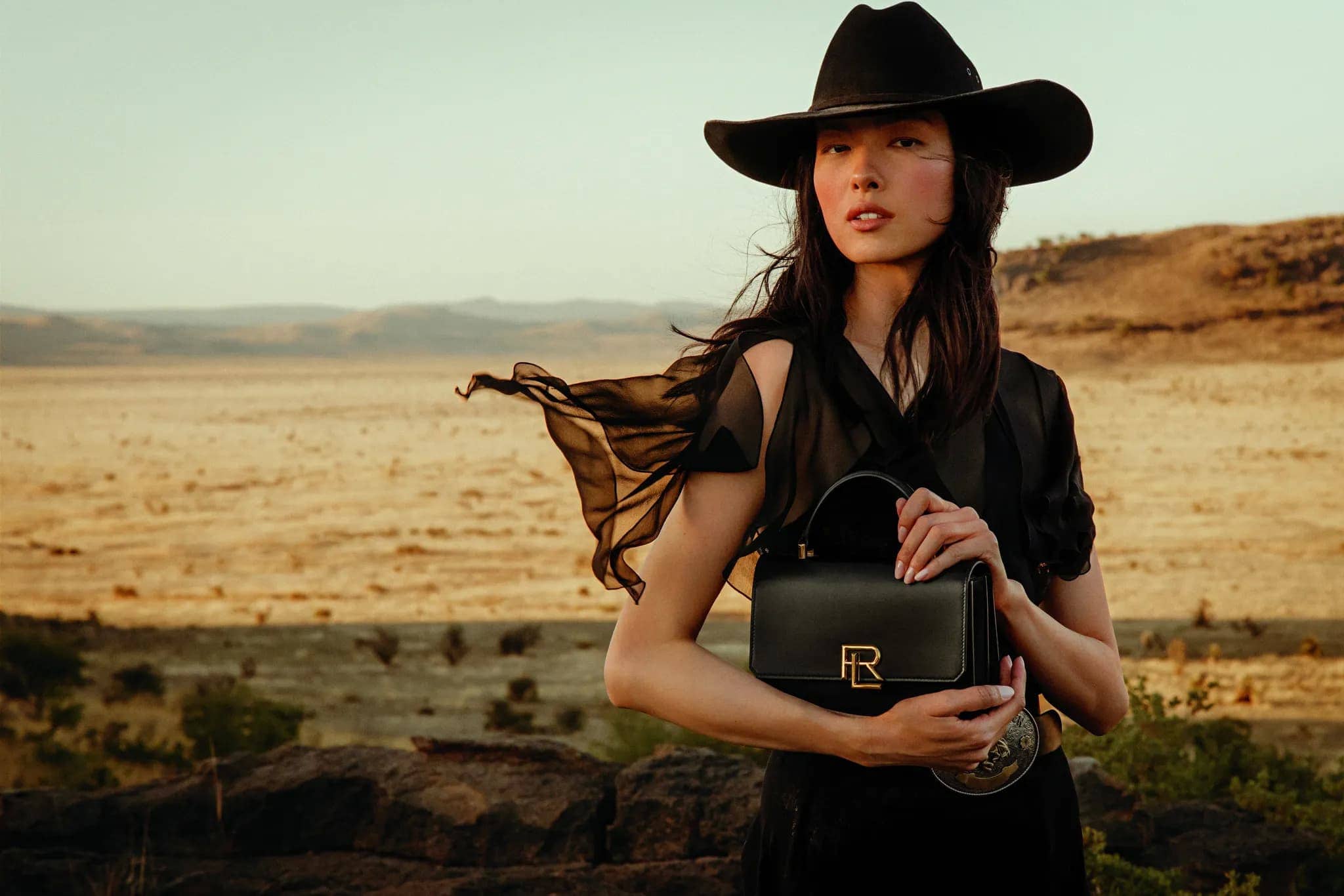 Woman in a black cowboy hat and dress holding a black leather purse, posing in a desert landscape with hills under a bright sky.