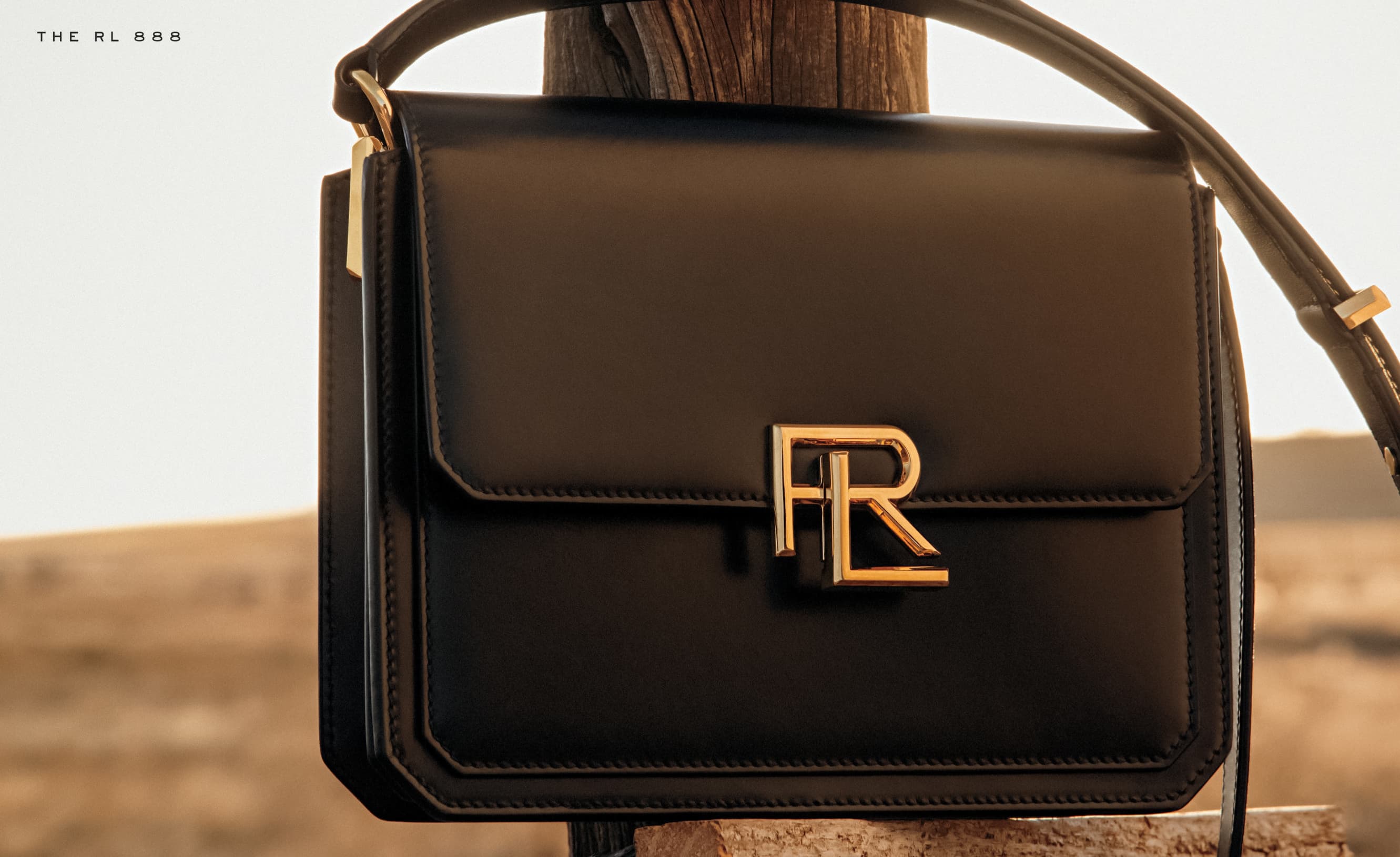 Elegant black leather shoulder bag with a gold logo clasp, set against a warm, blurred desert backdrop, creating a sophisticated fashion shot.