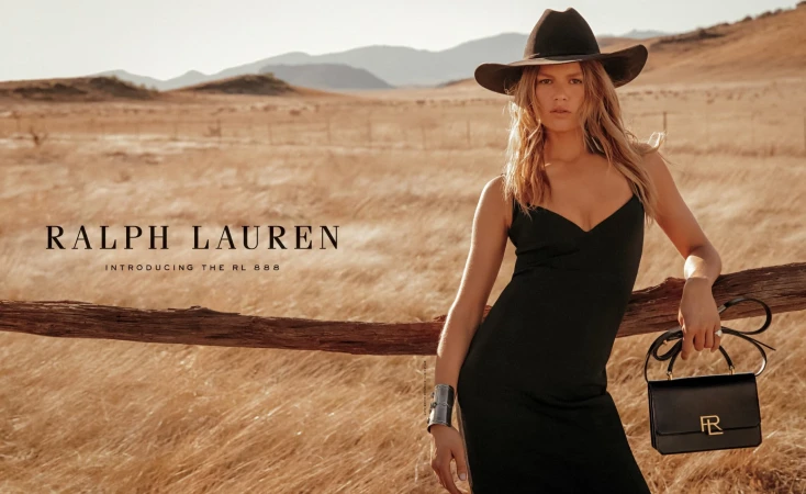 Fashion shot of a woman in a black dress and cowboy hat posing in front of a rural landscape. She carries a black leather handbag.