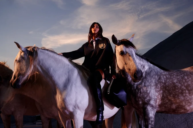 A woman in a black sweater and boots sits on a white horse amidst a group of horses, set against a dusky sky and dark landscape.