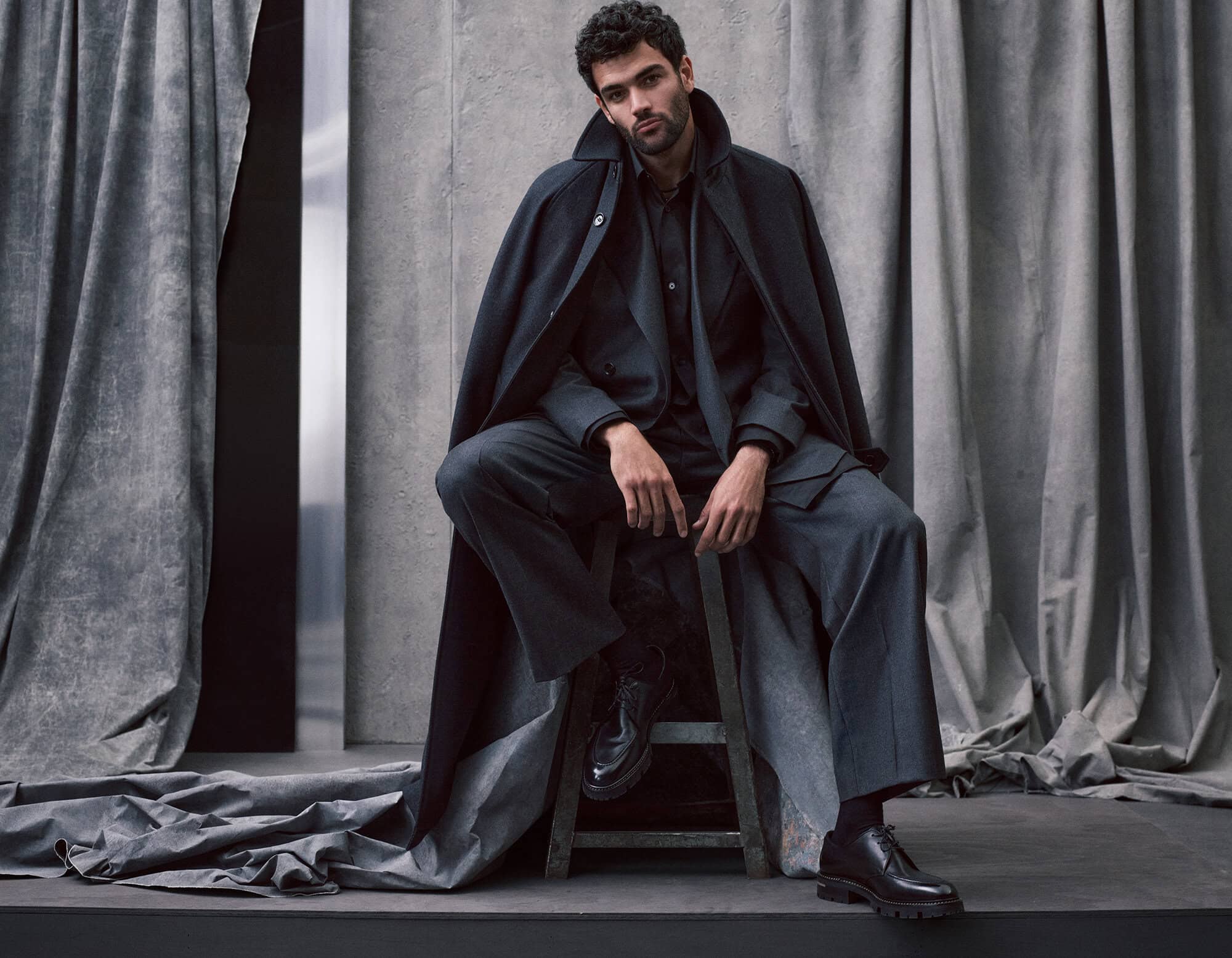 Man in a dark suit and cape sits on a stool in a studio with draped curtains. Fashion editorial shot with neutral color grading.
