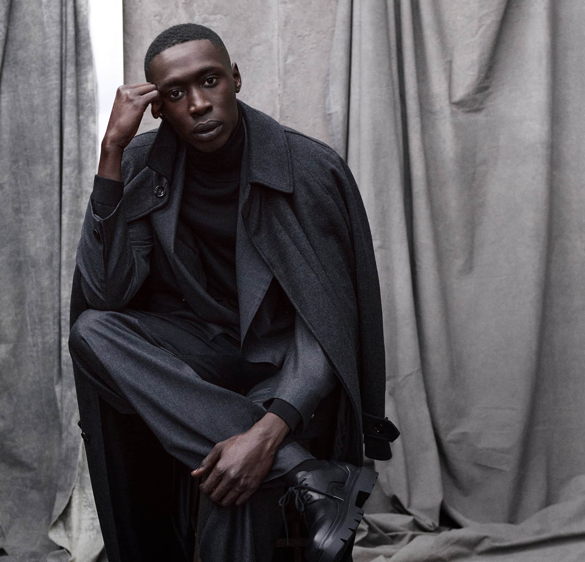 An African-American man is posing in a studio, wearing a grey suit and coat, with a black turtleneck, looking directly into the camera.