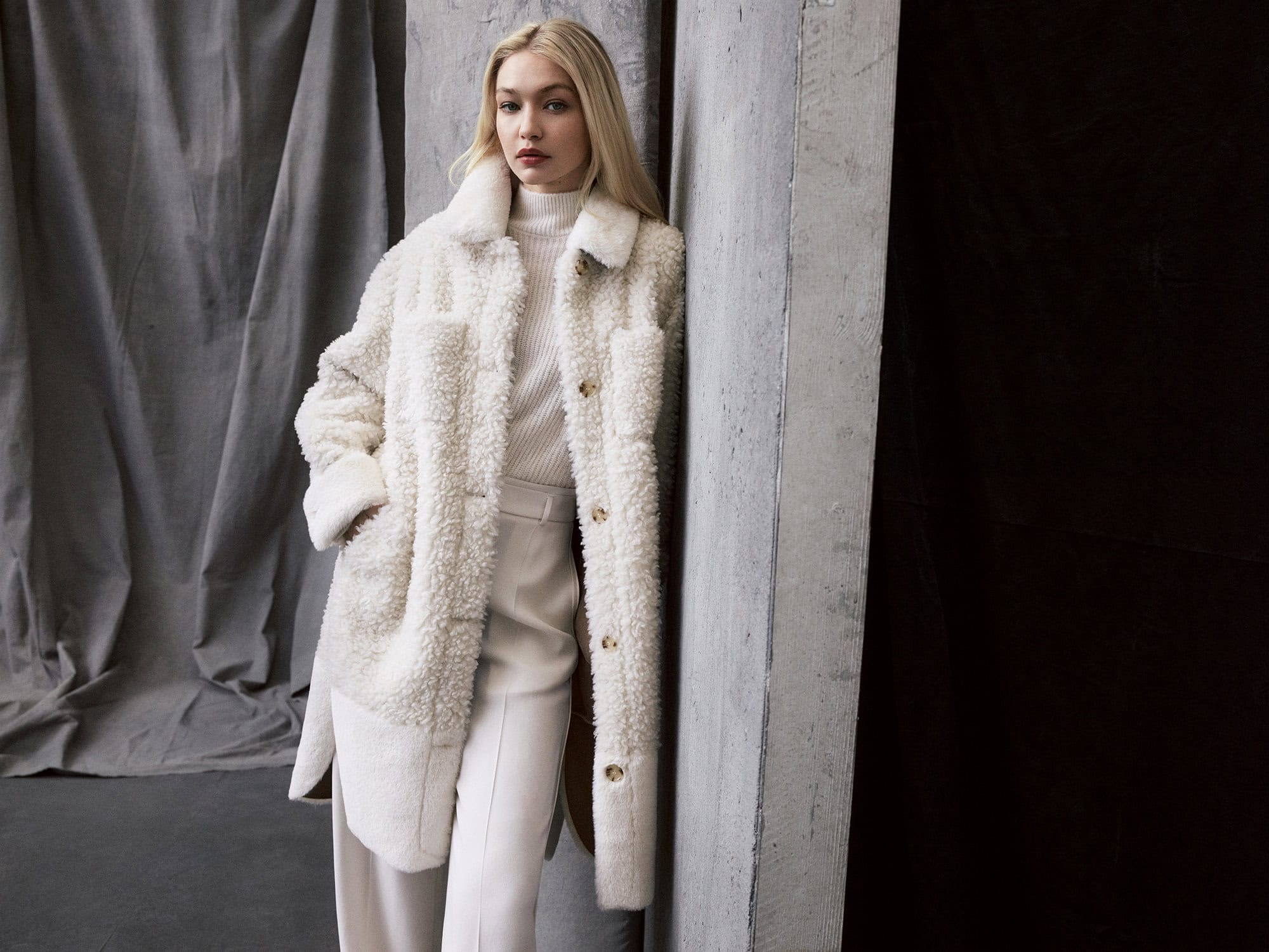 Fashion shot of a blonde female model posing indoors, wearing an off-white sherpa jacket, turtleneck and matching pants against a gray backdrop.