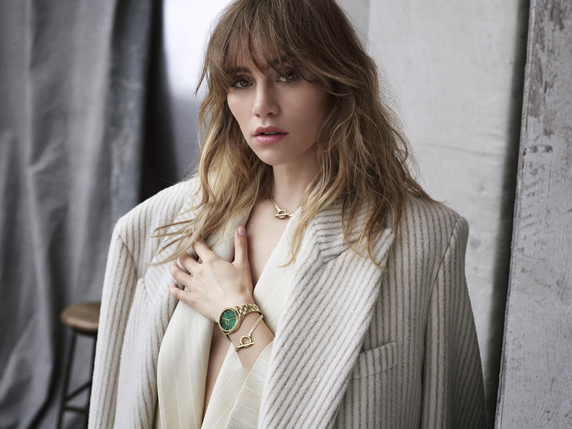 Close-up of a blonde woman in a pinstripe blazer and gold jewelry with a serious facial expression in an indoor environment.