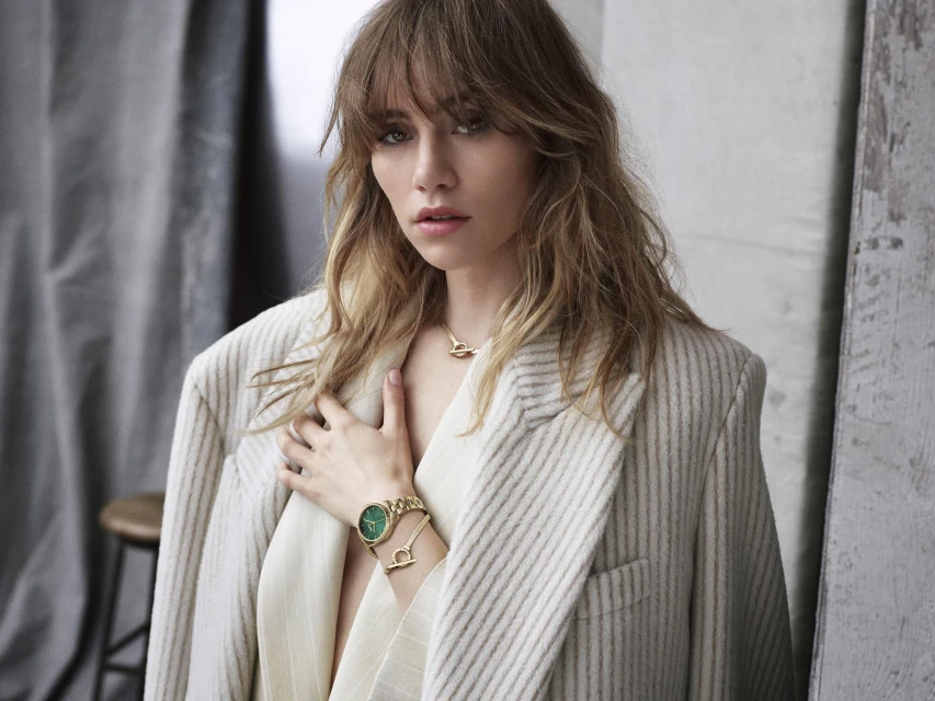 Close-up of a blonde woman in a pinstripe blazer and gold jewelry with a serious facial expression in an indoor environment.