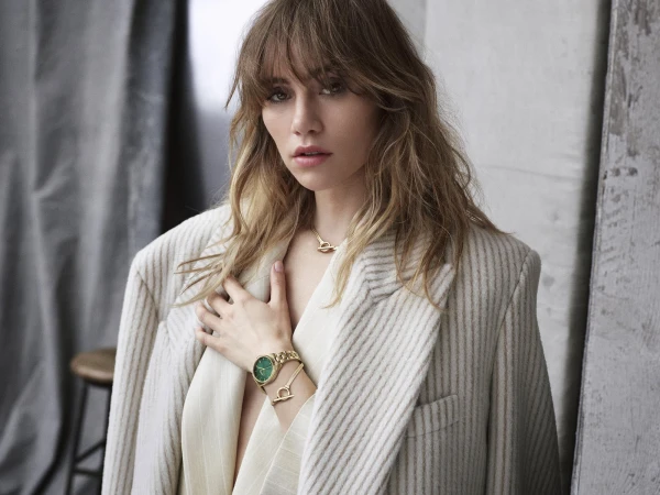 Close-up of a blonde woman in a pinstripe blazer and gold jewelry with a serious facial expression in an indoor environment.