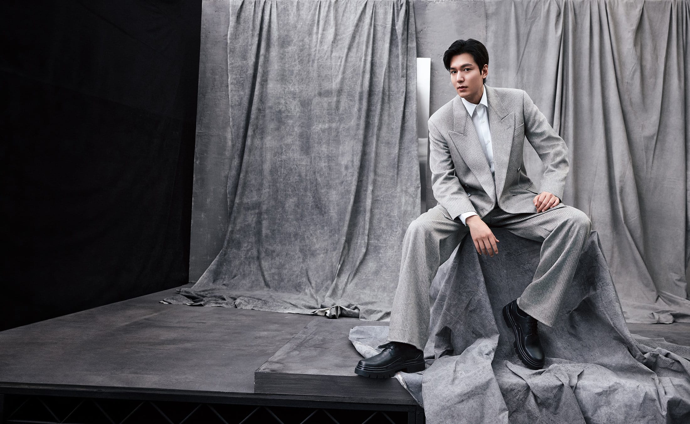 Man in grey suit seated on a platform, posing in a photo studio against a grey textured backdrop, looking at the camera with a neutral expression.