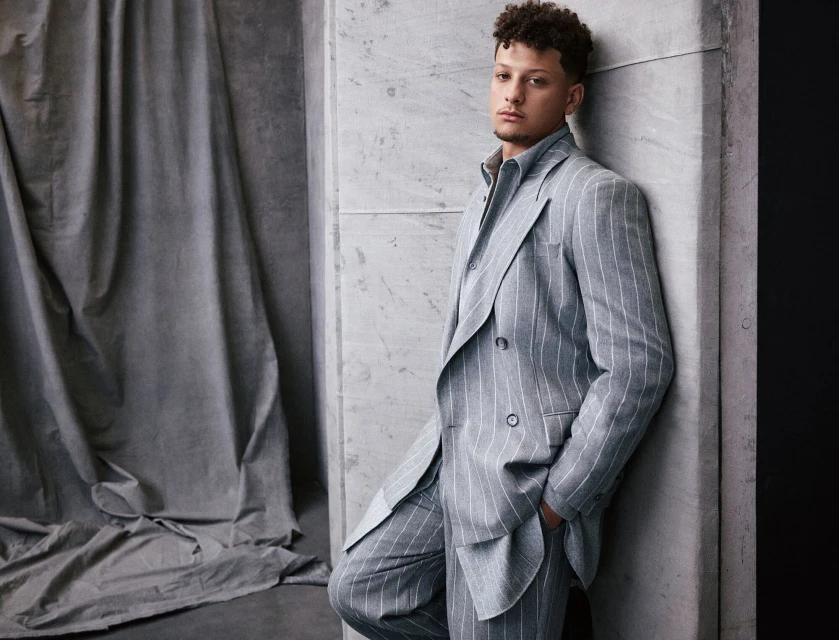 Adult male in a double-breasted striped suit leans against a concrete wall in an indoor photo studio setting with draped fabric.