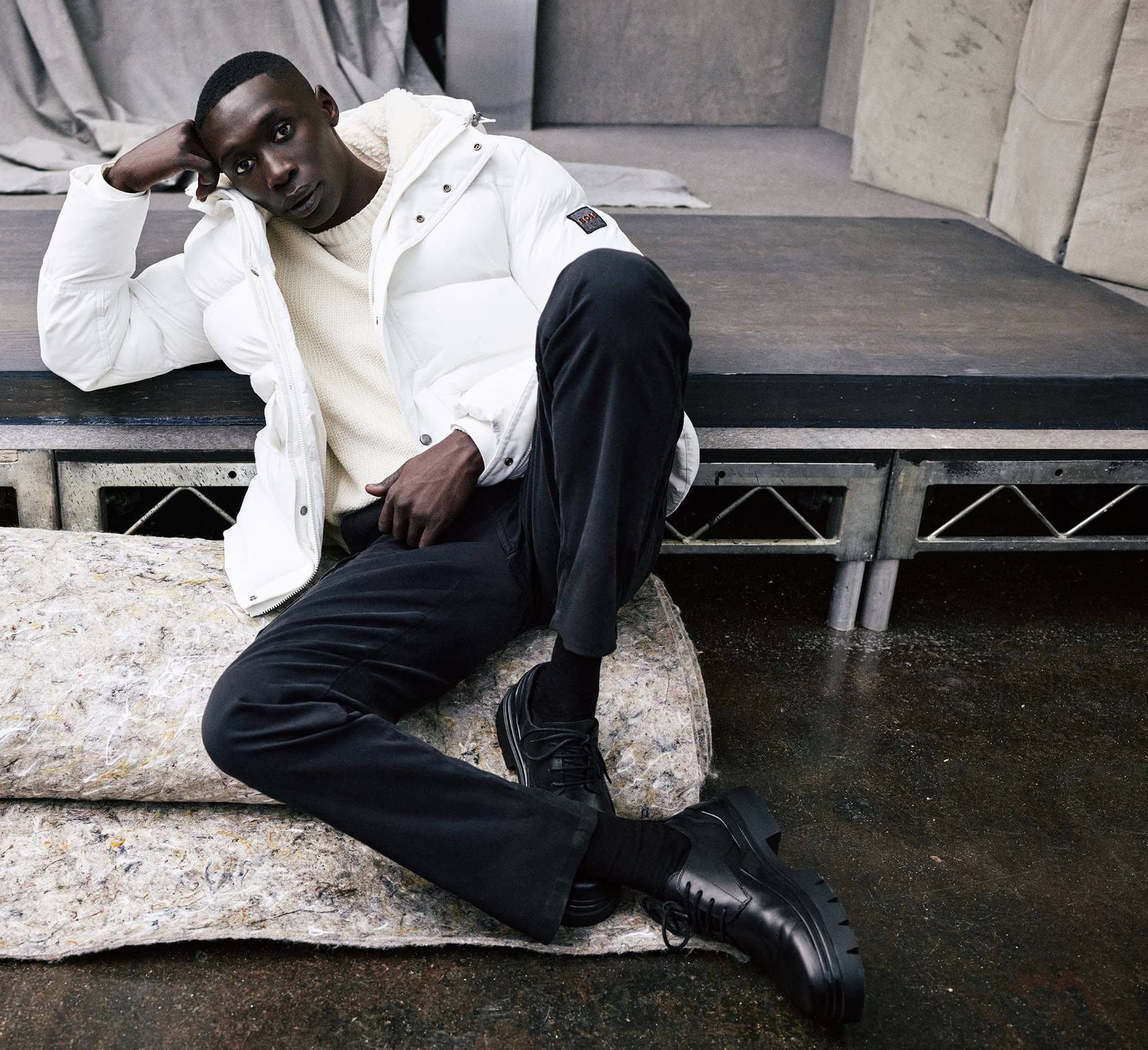 Man in white puffer jacket and black pants sitting on a textured platform with dark hair and complexion in a casual pose.