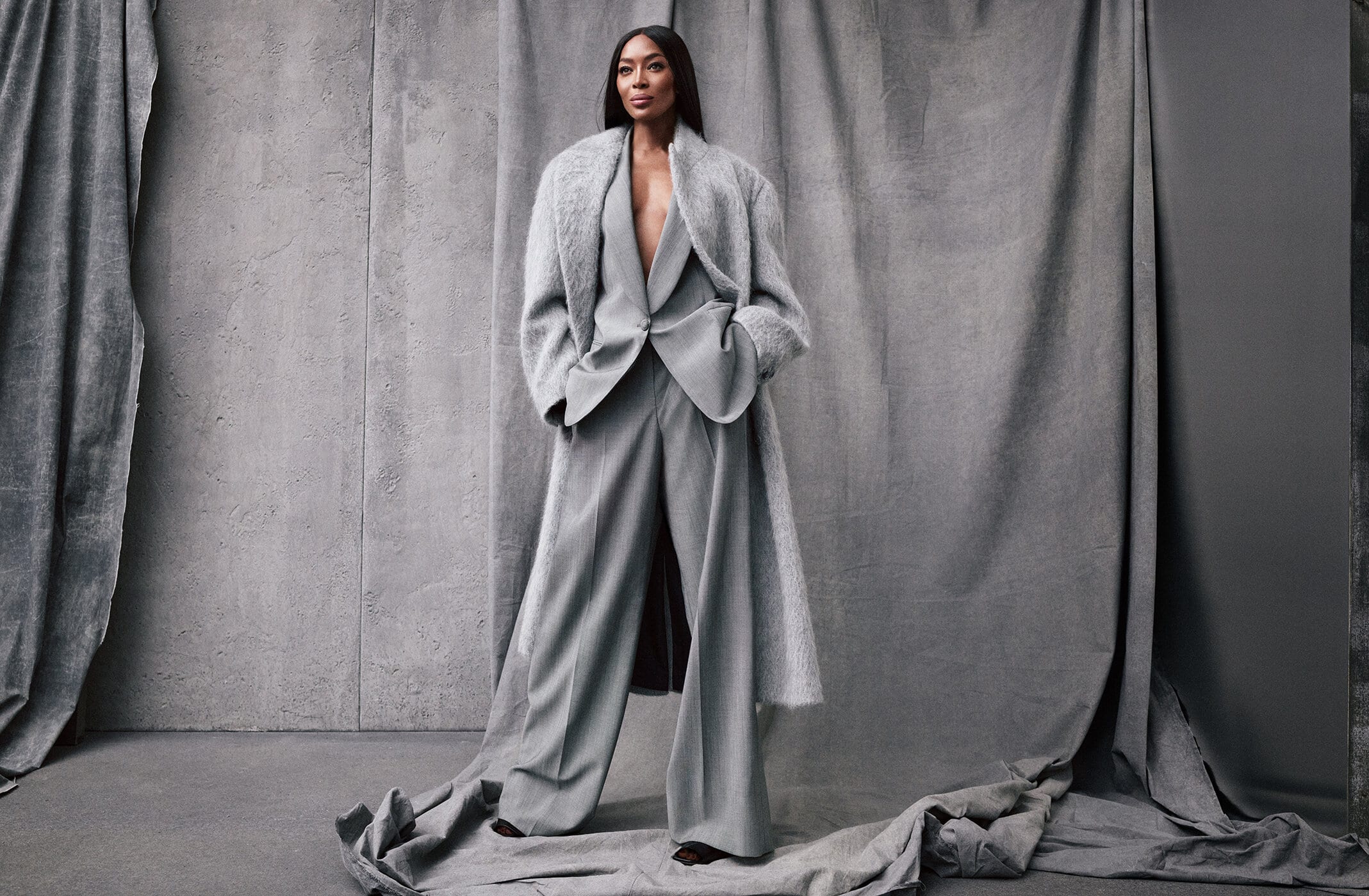 Naomi Campbell poses in a grey pantsuit and coat against a matching draped background in a studio portrait with soft lighting.