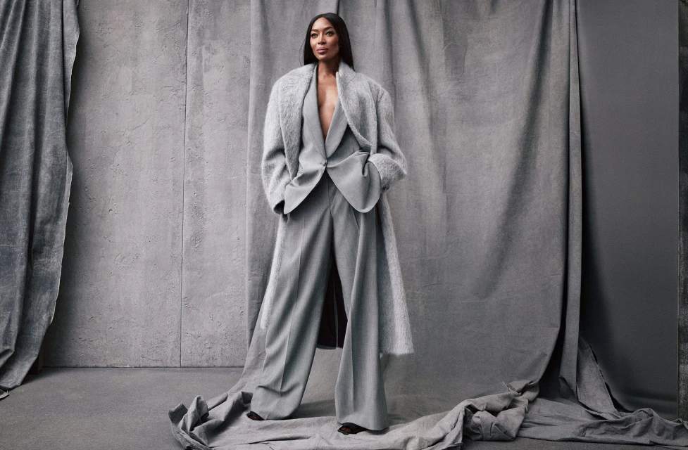 Naomi Campbell poses in a grey pantsuit and coat against a matching draped background in a studio portrait with soft lighting.