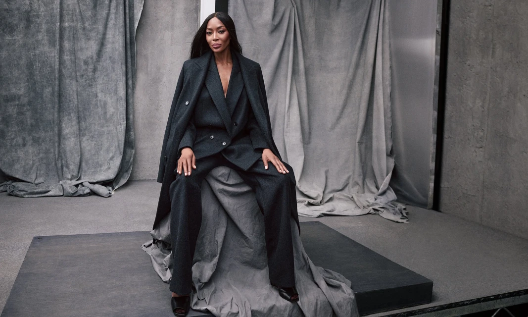 An adult female model sits on a gray surface, wearing a dark pantsuit and open overcoat in a studio setting with draped gray fabric.