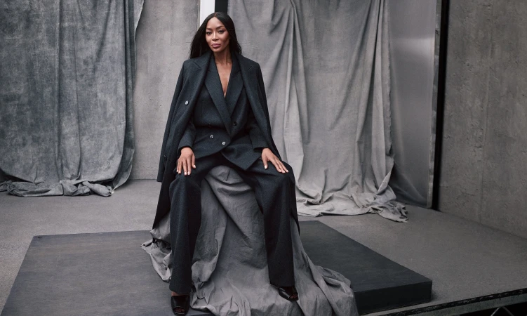 An adult female model sits on a gray surface, wearing a dark pantsuit and open overcoat in a studio setting with draped gray fabric.