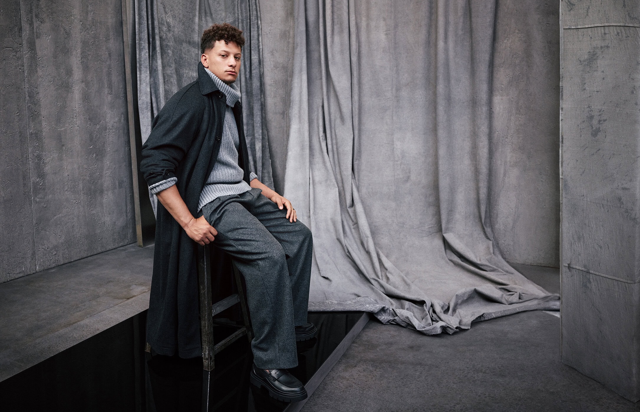 Adult male poses in studio wearing a grey turtleneck sweater, trousers, black overcoat, and black shoes. He is sitting on a chair.