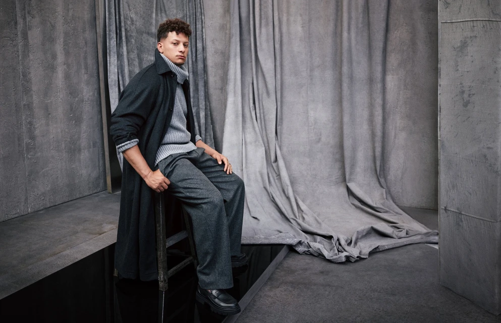 Adult male poses in studio wearing a grey turtleneck sweater, trousers, black overcoat, and black shoes. He is sitting on a chair.