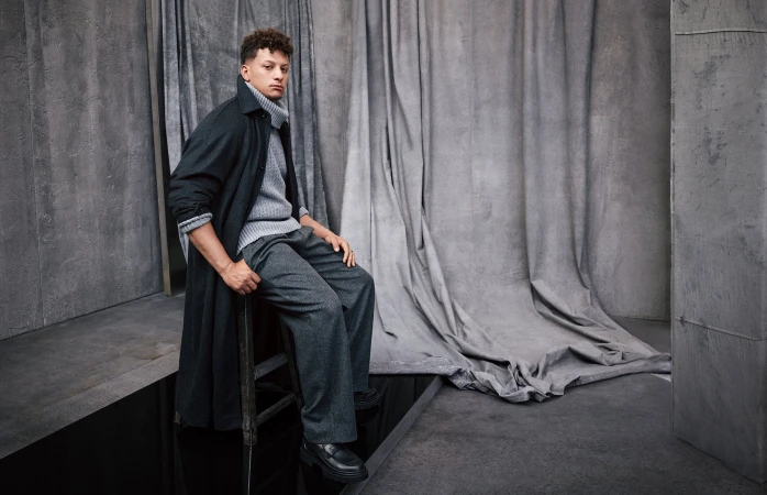 Adult male poses in studio wearing a grey turtleneck sweater, trousers, black overcoat, and black shoes. He is sitting on a chair.
