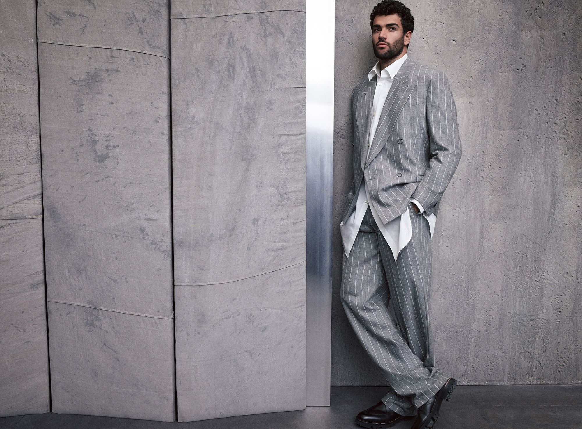 An adult male model with curly dark hair, wearing a gray pinstripe suit, poses in a photo studio against a gray textured wall.