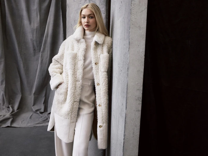 Gigi Hadid leaning against a concrete pillar wearing a white textured fleece coat and matching trousers for BOSS Fall 2023.