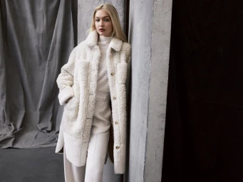Gigi Hadid leaning against a concrete pillar wearing a white textured fleece coat and matching trousers for BOSS Fall 2023.