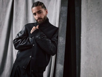 Maluma wearing a black bomber jacket with patterned sleeves leaning against a grey concrete pillar in a studio portrait.