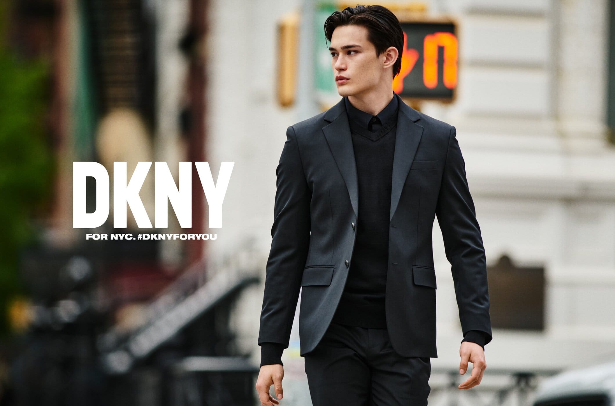 Handsome man in DKNY suit walks in urban setting. Stylish menswear fashion advertisement on city street with blurred background.
