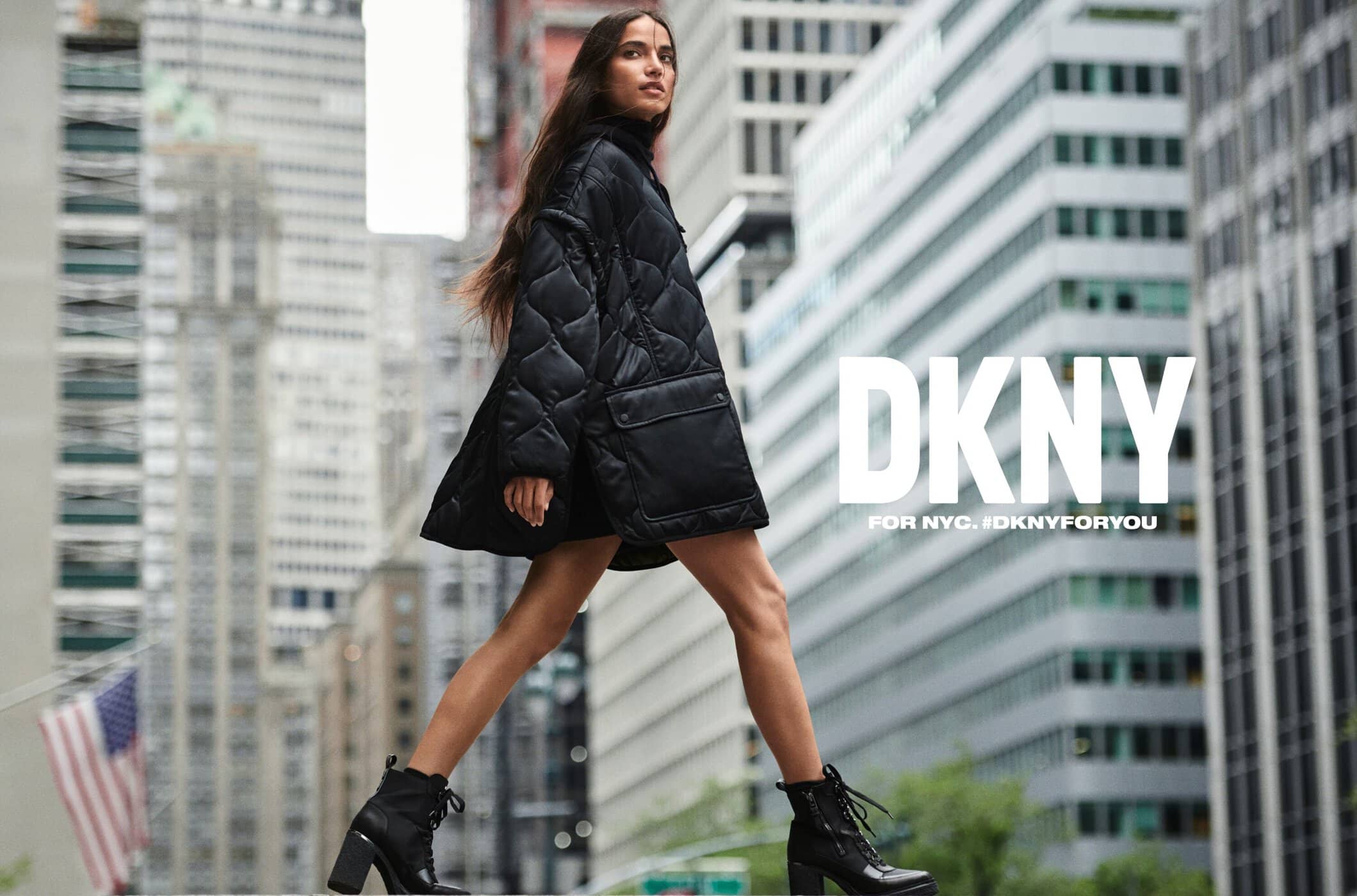 Fashion model walking in city wearing black quilted jacket and boots with DKNY advertisement in the background, urban fashion advertisement.