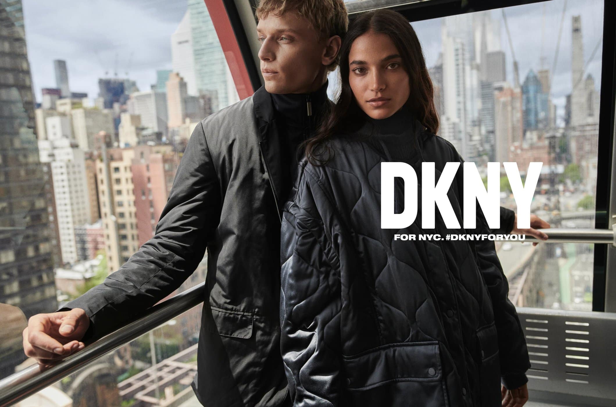 Fashionable couple in black coats stands in modern cable car against a city skyline. DKNY brand visible, promoting New York City style.