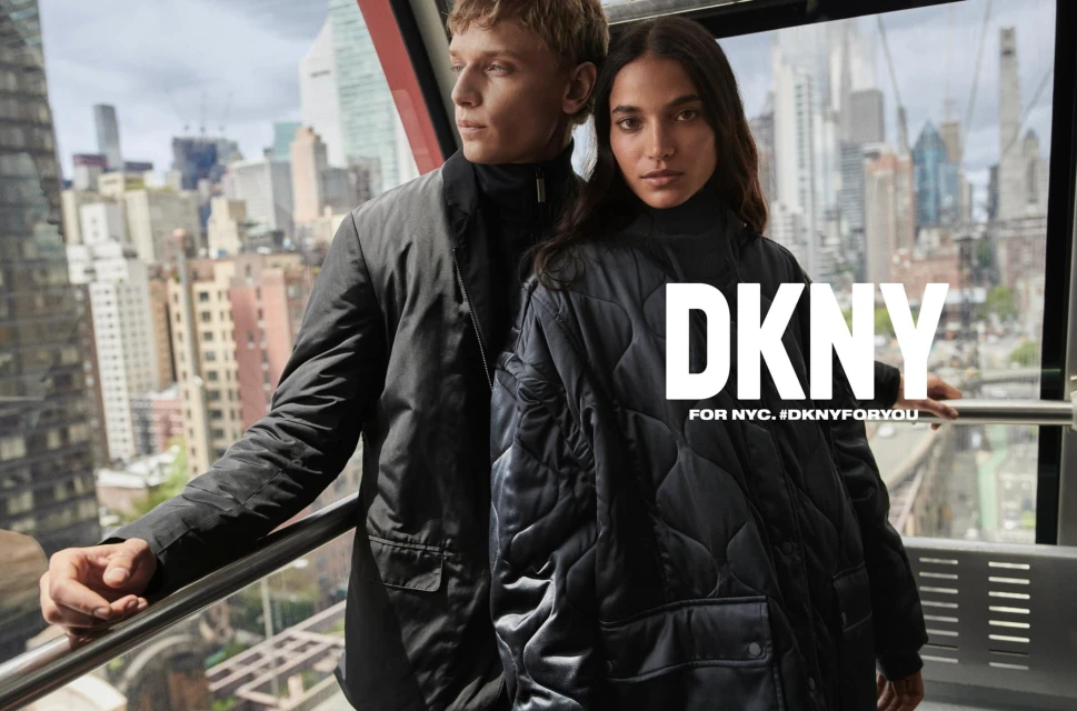 Fashionable couple in black coats stands in modern cable car against a city skyline. DKNY brand visible, promoting New York City style.