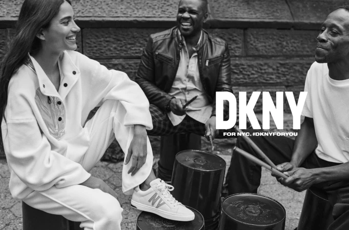 Black and white fashion ad featuring a smiling woman in white loungewear, drummers, and DKNY logo against an urban backdrop.