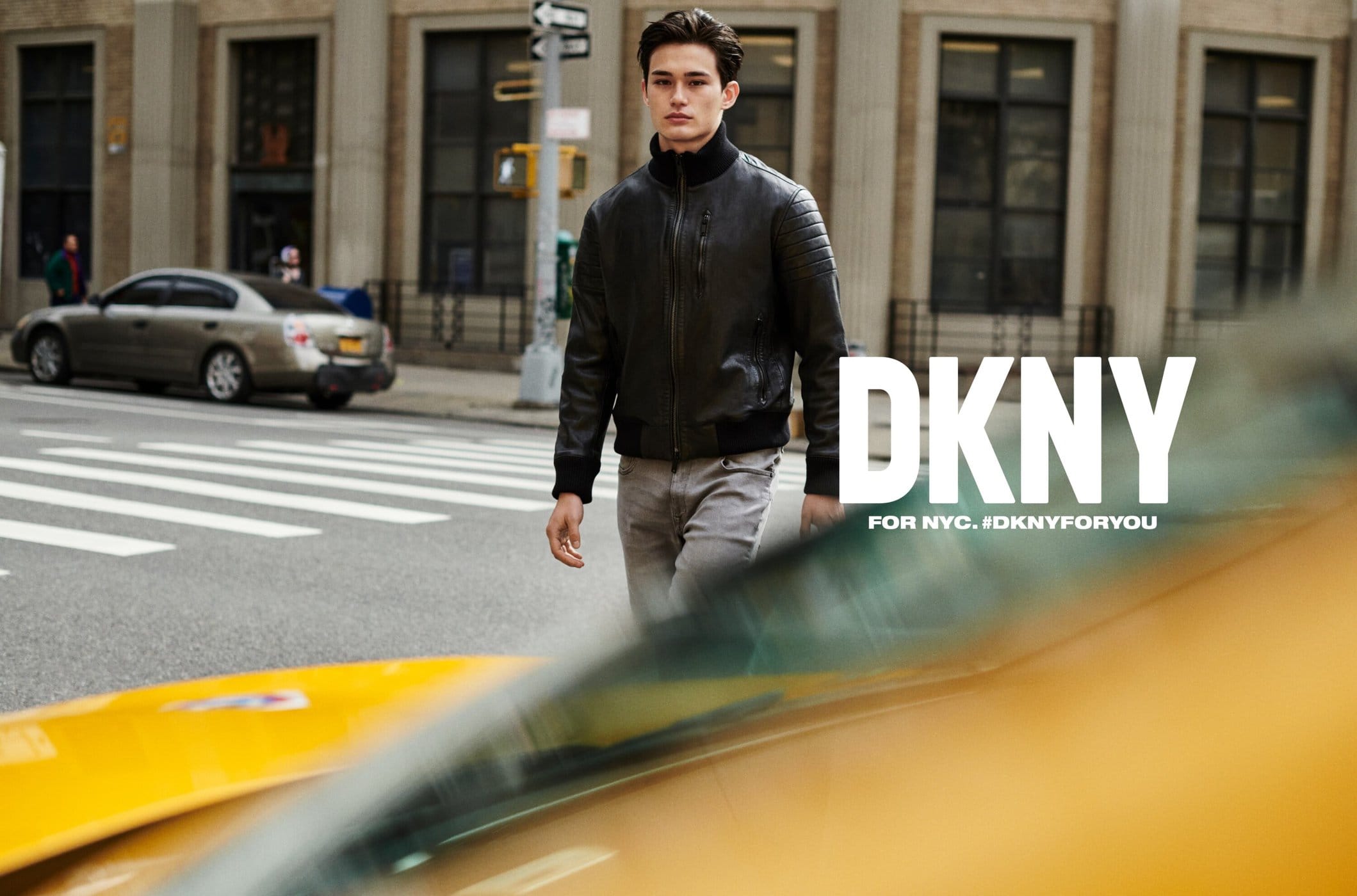 A young adult male is walking on a city street wearing a black jacket, grey pants, and black wrist cuffs with a DKNY advert in frame.
