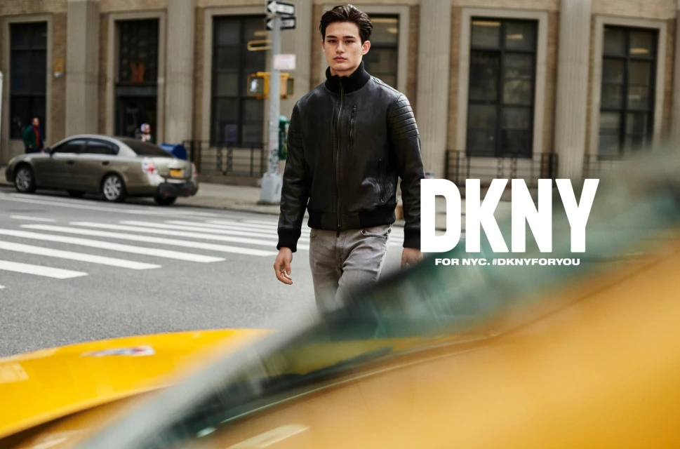 A young adult male is walking on a city street wearing a black jacket, grey pants, and black wrist cuffs with a DKNY advert in frame.