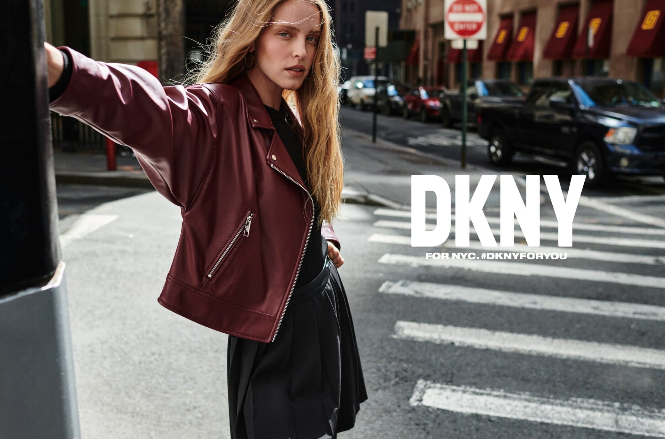 Blonde woman in a burgundy jacket stands on a city street with cars and buildings in the background and DKNY branding.