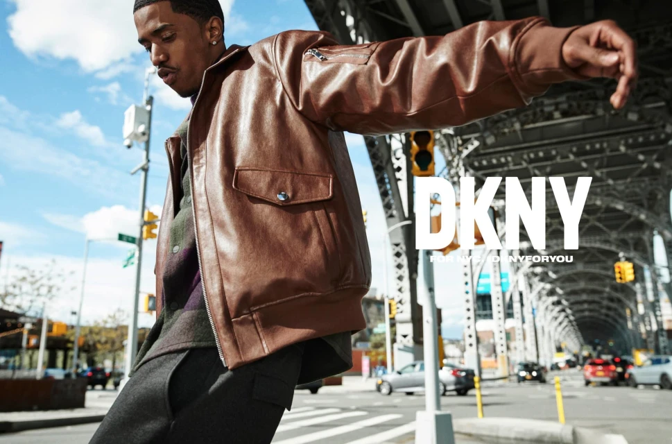 Outdoor fashion shoot of a young adult male model in a brown leather jacket standing on a city street in front of a bridge with the DKNY logo.