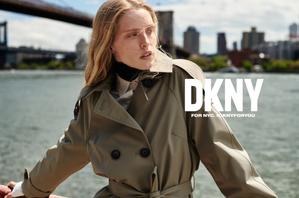 Fashion advertisement featuring a blonde woman in a trench coat, with a city skyline and bridge in the background and DKNY logo.