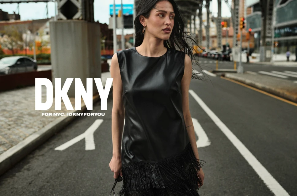A woman in a black dress poses on a city street with a bridge in the background for a DKNY fashion advertisement campaign.
