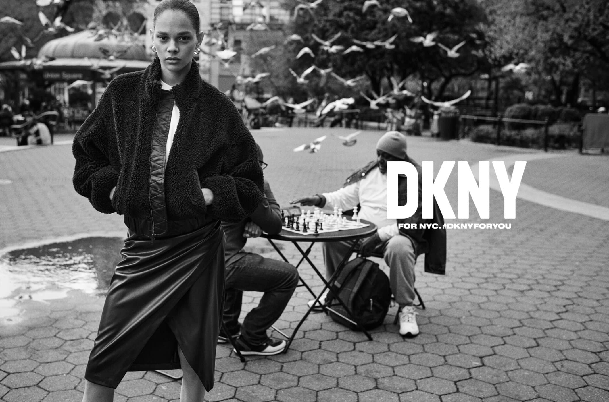 Fashion shot in a city park with a woman in a black jacket and skirt, people playing chess, and pigeons flying overhead in black and white.