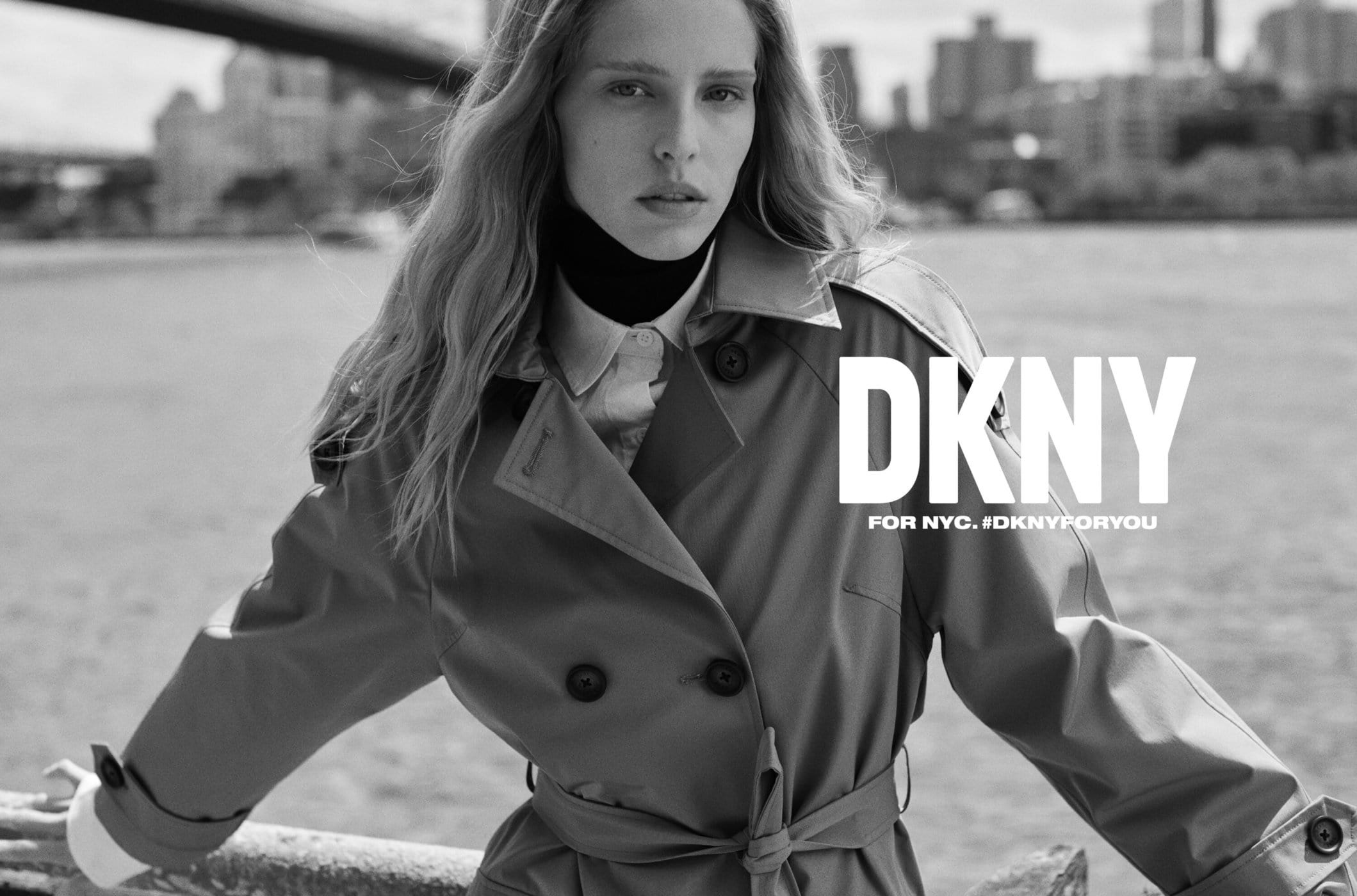 Black and white fashion portrait of an adult female model wearing a trench coat with a river and cityscape background.