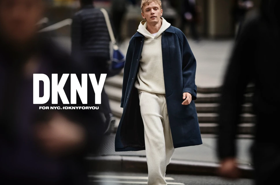 Fashion advertisement featuring a male model in a navy coat and cream-colored sweatsuit walking on a city street with blurred pedestrians.