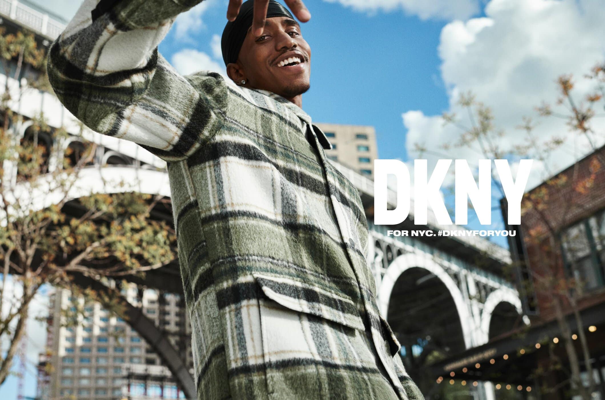 Young man smiles wearing a green and white plaid flannel jacket outdoors with city buildings, blue sky, and DKNY logo in the background.